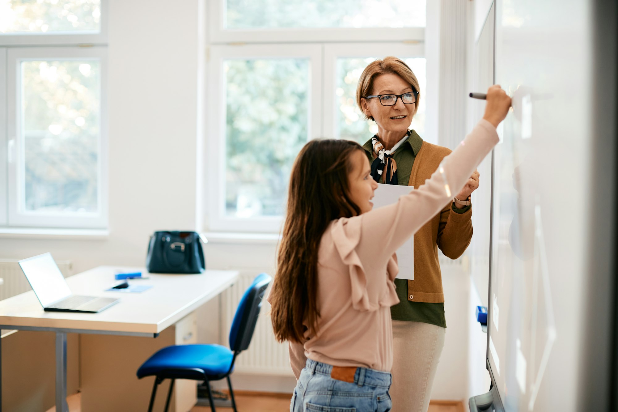 Mature teacher assisting schoolgirl in writing assignment on smartboard in the classroom.